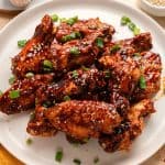 A plate of glazed chicken wings garnished with sesame seeds and chopped green onions, with garlic bulbs and a bowl of sesame seeds in the background.