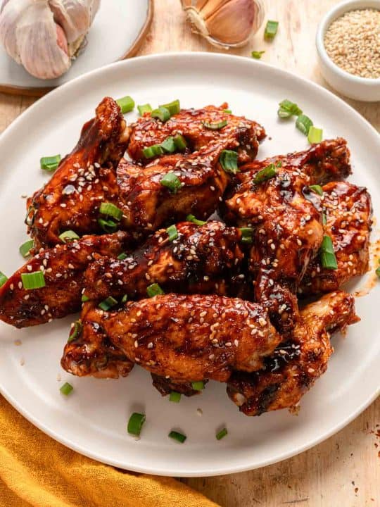 A plate of glazed chicken wings garnished with sesame seeds and chopped green onions, with garlic bulbs and a bowl of sesame seeds in the background.