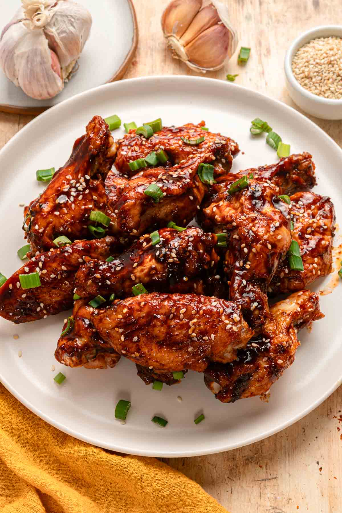 A plate of glazed chicken wings garnished with sesame seeds and chopped green onions, with garlic bulbs and a bowl of sesame seeds in the background.