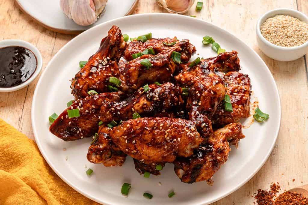 A plate of glazed chicken wings garnished with sesame seeds and chopped green onions, set on a wooden table with sauces and seasonings nearby.