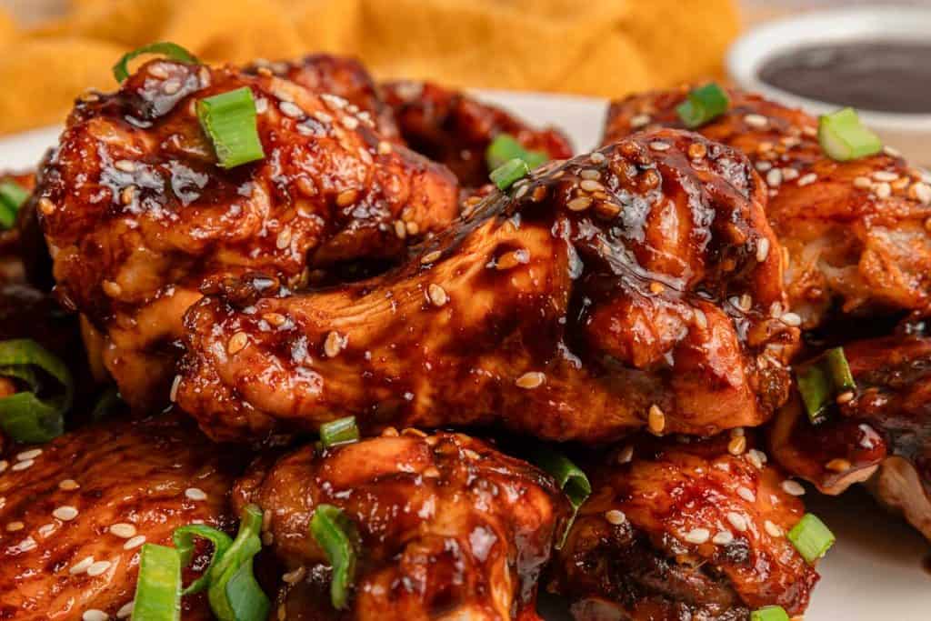 A close-up of glazed chicken wings garnished with sesame seeds and sliced green onions, with a small dish of sauce in the background.