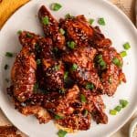 A plate of glazed chicken wings garnished with sesame seeds and chopped green onions, placed on a wooden table with a yellow cloth and garlic nearby.