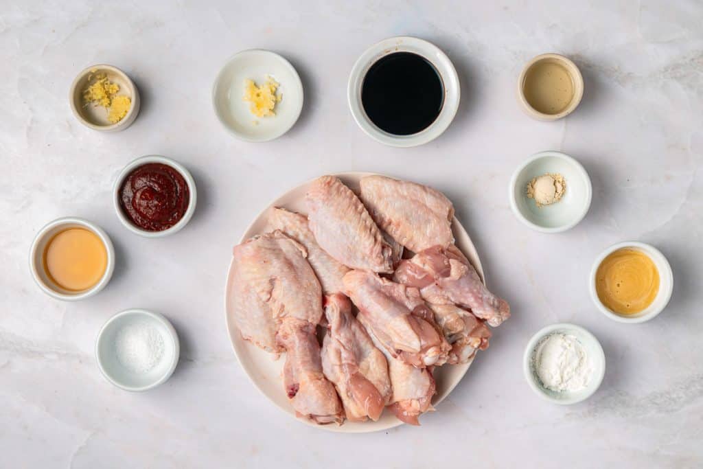 A plate of raw chicken drumsticks and wings is surrounded by small bowls containing various sauces, spices, and seasonings on a light countertop.