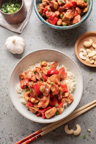 A bowl of rice topped with stir-fried chicken, red bell peppers, and cashews next to chopsticks, a bowl of green onions, raw cashews, and a garlic bulb on a gray surface.