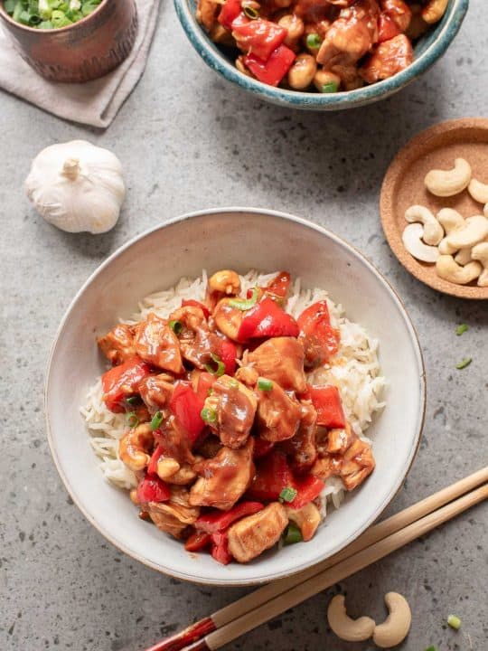 A bowl of rice topped with stir-fried chicken, red bell peppers, and cashews next to chopsticks, a bowl of green onions, raw cashews, and a garlic bulb on a gray surface.