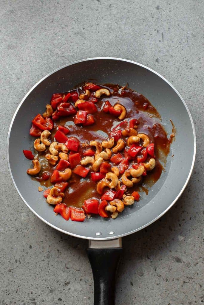 A frying pan on a gray countertop contains red bell peppers, cashews, and a brown sauce.