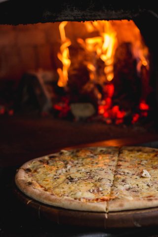 A cheese pizza with herbs sits in front of a wood-fired oven with burning flames and glowing embers.
