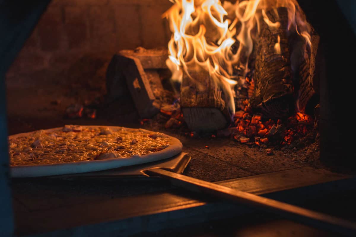 A pizza on a wooden peel being placed into a wood-fired oven with flames and burning logs visible.