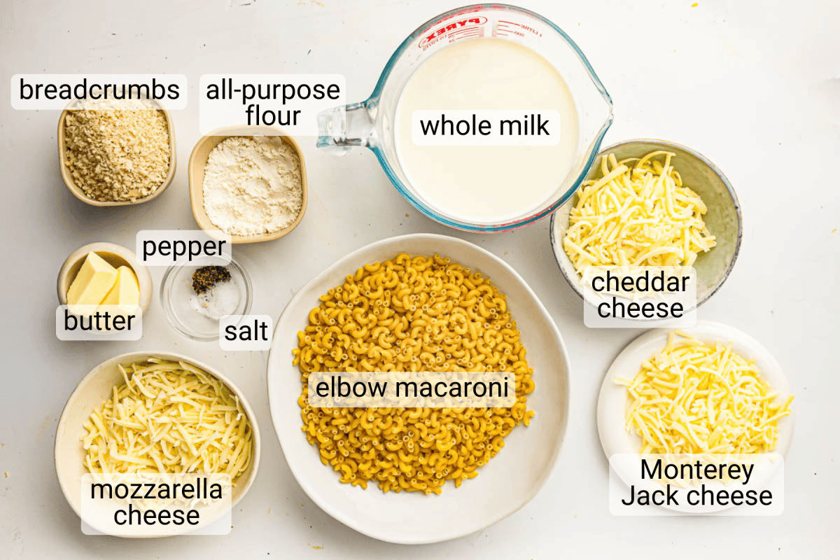 Various ingredients for macaroni and cheese are arranged on a white surface, each labeled, including macaroni, cheeses, whole milk, flour, butter, breadcrumbs, salt, and pepper.