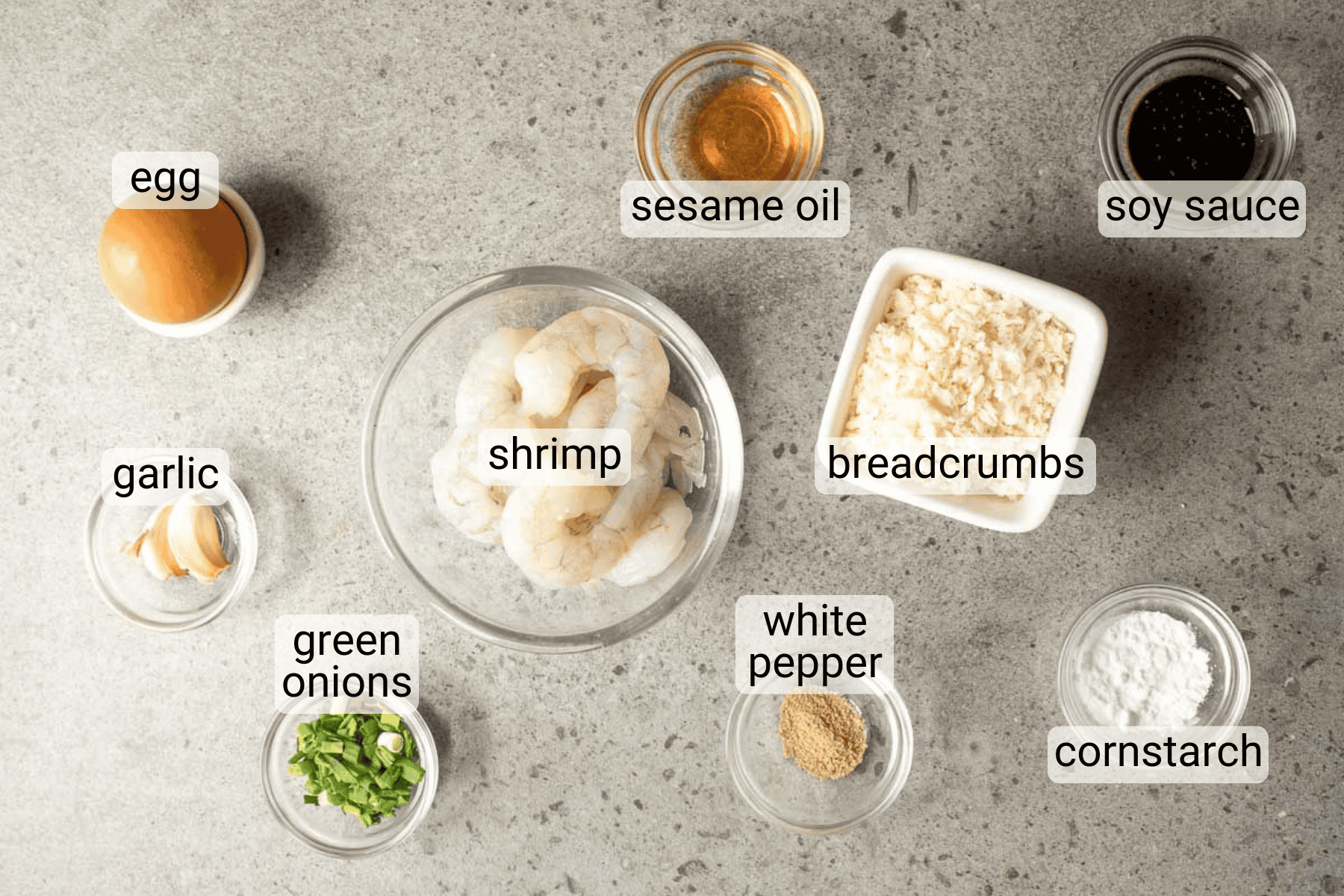 A flat lay of shrimp, egg, breadcrumbs, green onions, garlic, white pepper, cornstarch, soy sauce, and sesame oil in separate containers on a gray surface, each labeled.