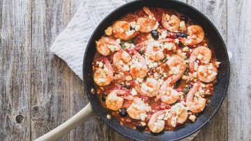 A skillet filled with shrimp in a tomato-based sauce, topped with crumbled cheese, on a wooden table beside a folded cloth.
