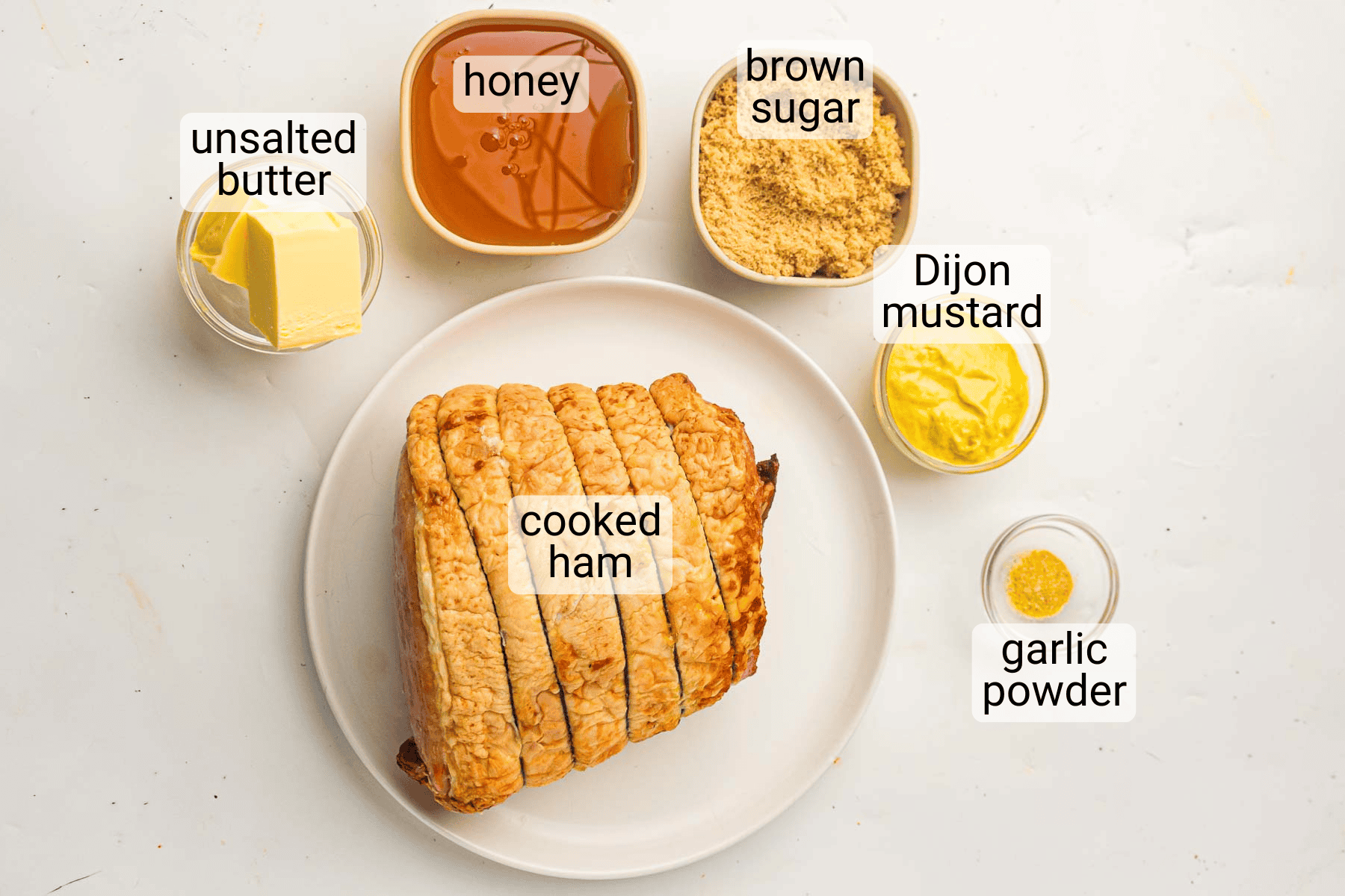 A plate with cooked ham in the center, surrounded by bowls containing unsalted butter, honey, brown sugar, Dijon mustard, and garlic powder, each labeled.