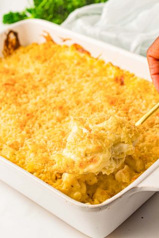 A hand scooping baked macaroni and cheese with a golden breadcrumb topping from a white casserole dish.