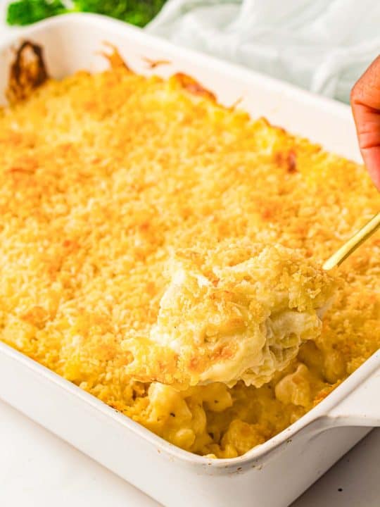 A hand scooping baked macaroni and cheese with a golden breadcrumb topping from a white casserole dish.