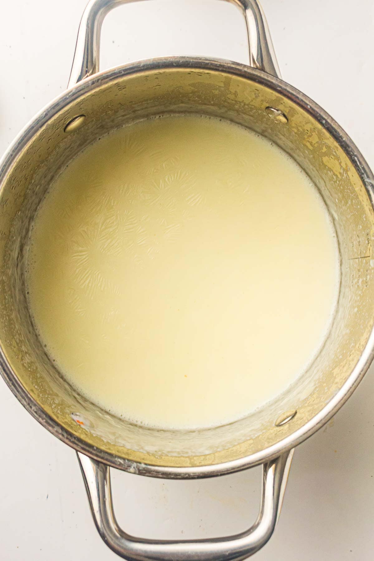 A stainless steel pot containing a creamy white liquid, likely milk or a milk-based mixture, sits on a white surface.