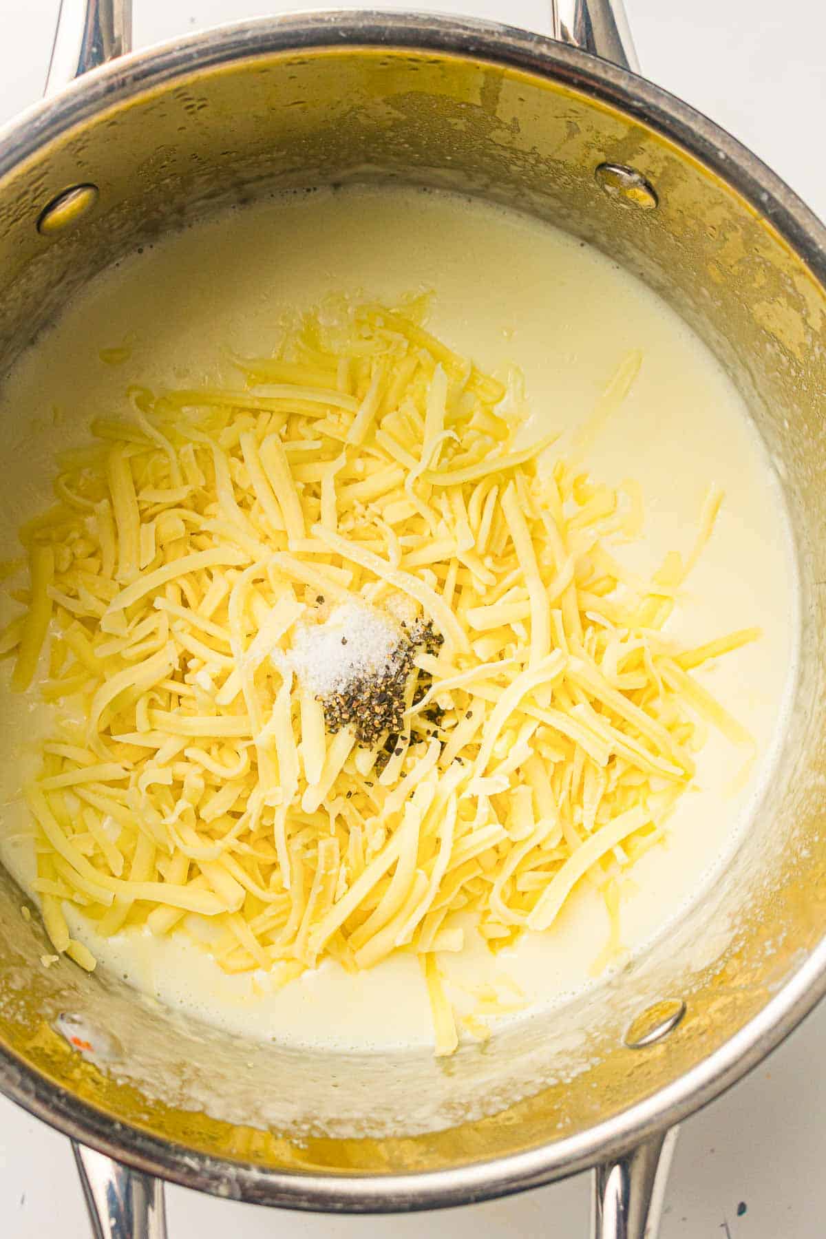 A metal saucepan containing shredded cheese, milk or cream, salt, and pepper, shown on a stovetop, ready to be mixed.