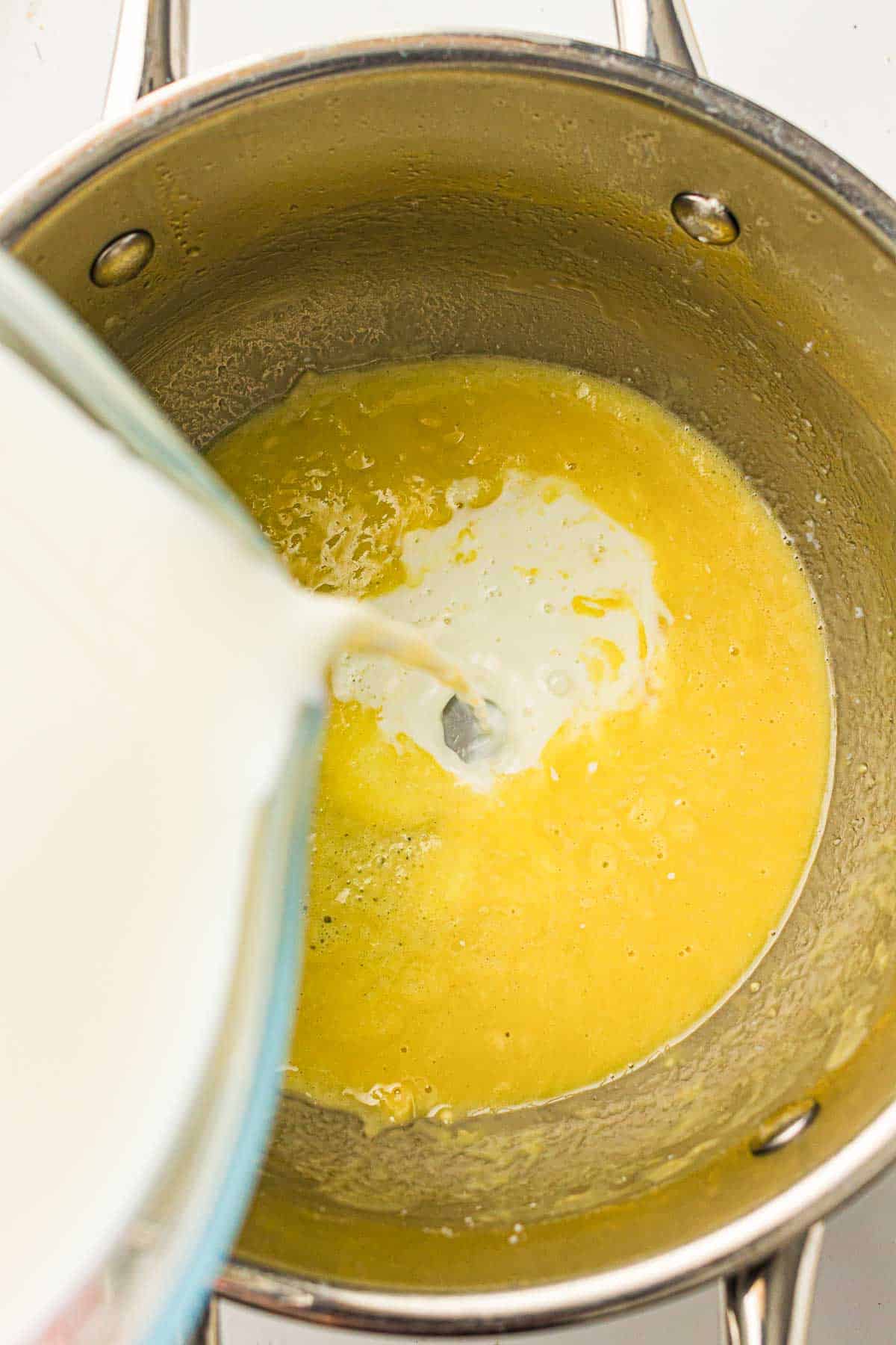 Milk is being poured from a glass container into a saucepan containing a yellow mixture, possibly eggs, on a stovetop.