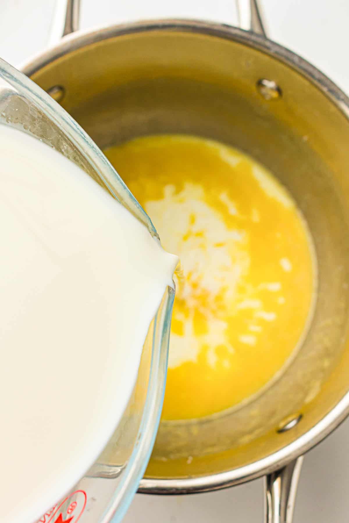 Milk is being poured from a glass measuring cup into a saucepan containing melted butter.