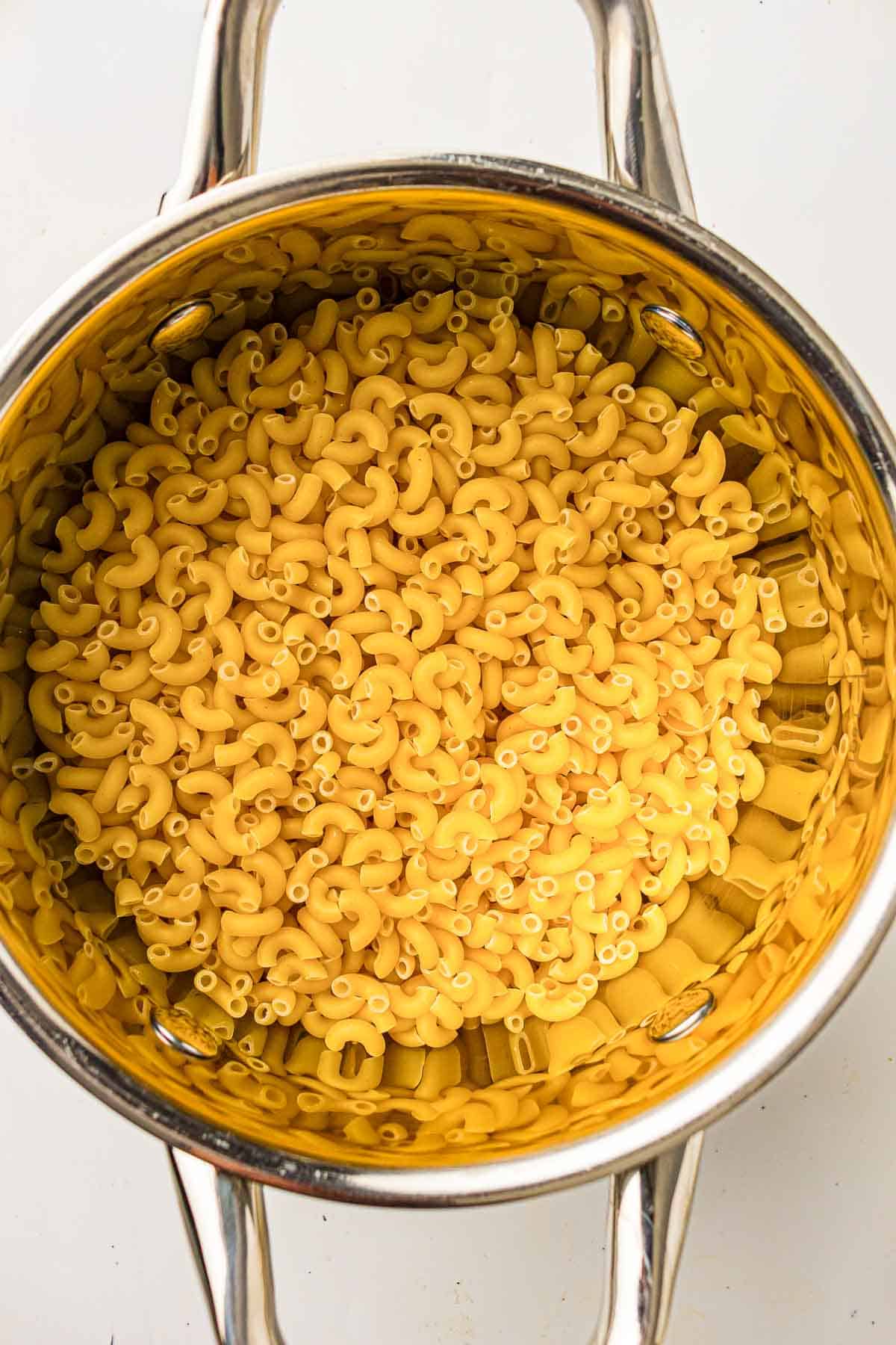 A stainless steel pot filled with uncooked elbow macaroni pasta viewed from above.