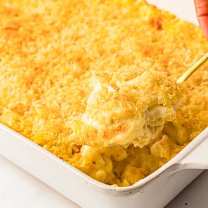 A hand scoops a serving of baked macaroni and cheese with a golden breadcrumb topping from a white casserole dish.