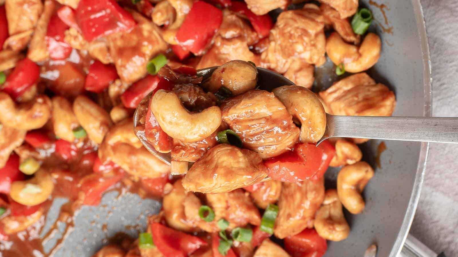 A spoonful of cashew chicken with red bell peppers and green onions held above a pan filled with the same dish.