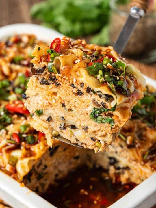 A close-up of a spatula lifting a slice of vegetable lasagna from a baking dish, showing layers of noodles, vegetables, sauce, and garnished with herbs and chili slices.