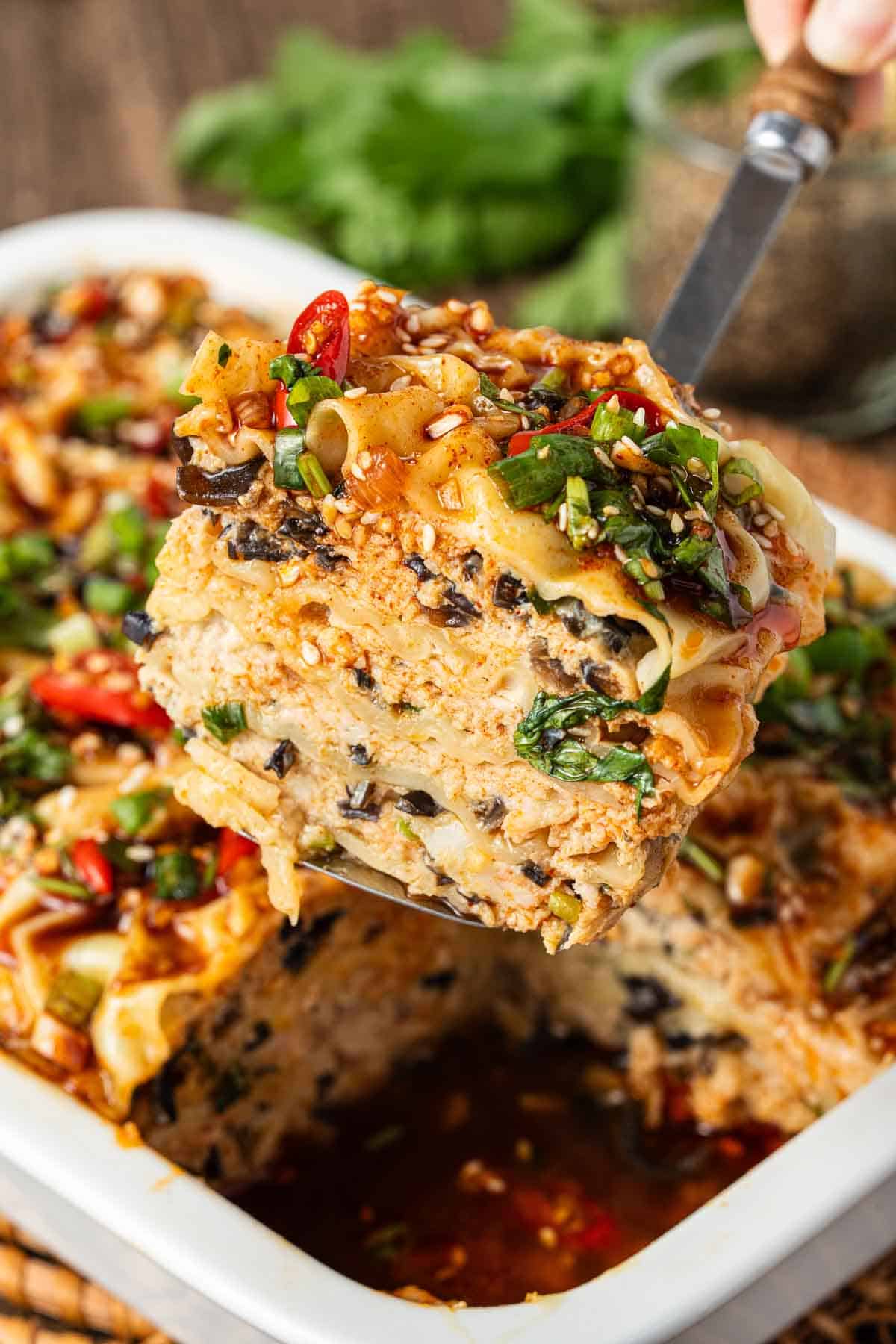 A close-up of a spatula lifting a slice of vegetable lasagna from a baking dish, showing layers of noodles, vegetables, sauce, and garnished with herbs and chili slices.