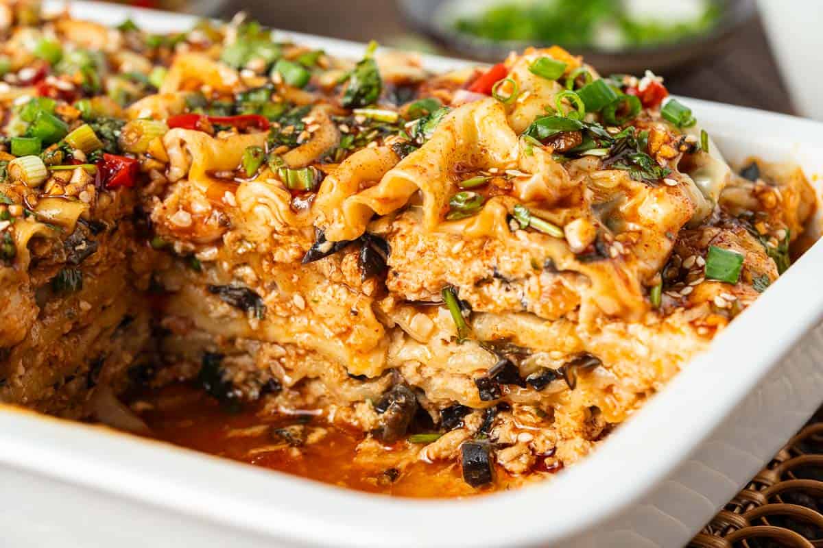 Close-up of a lasagna in a white baking dish, topped with chopped green onions and herbs, with a portion removed to show the layered interior.
