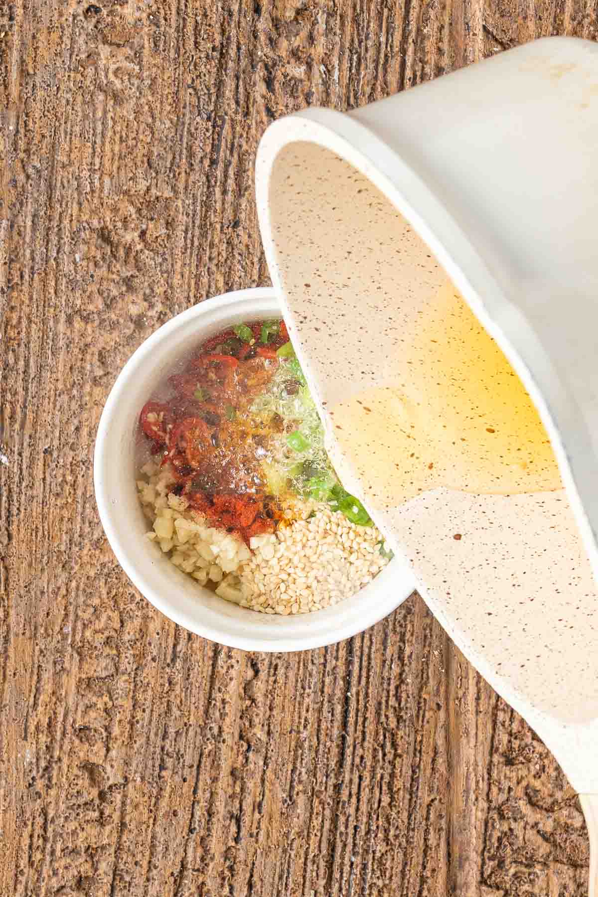 A white bowl with chopped garlic, sesame seeds, chopped green onions, and red spices being topped with hot oil from a tilted container on a wooden surface.