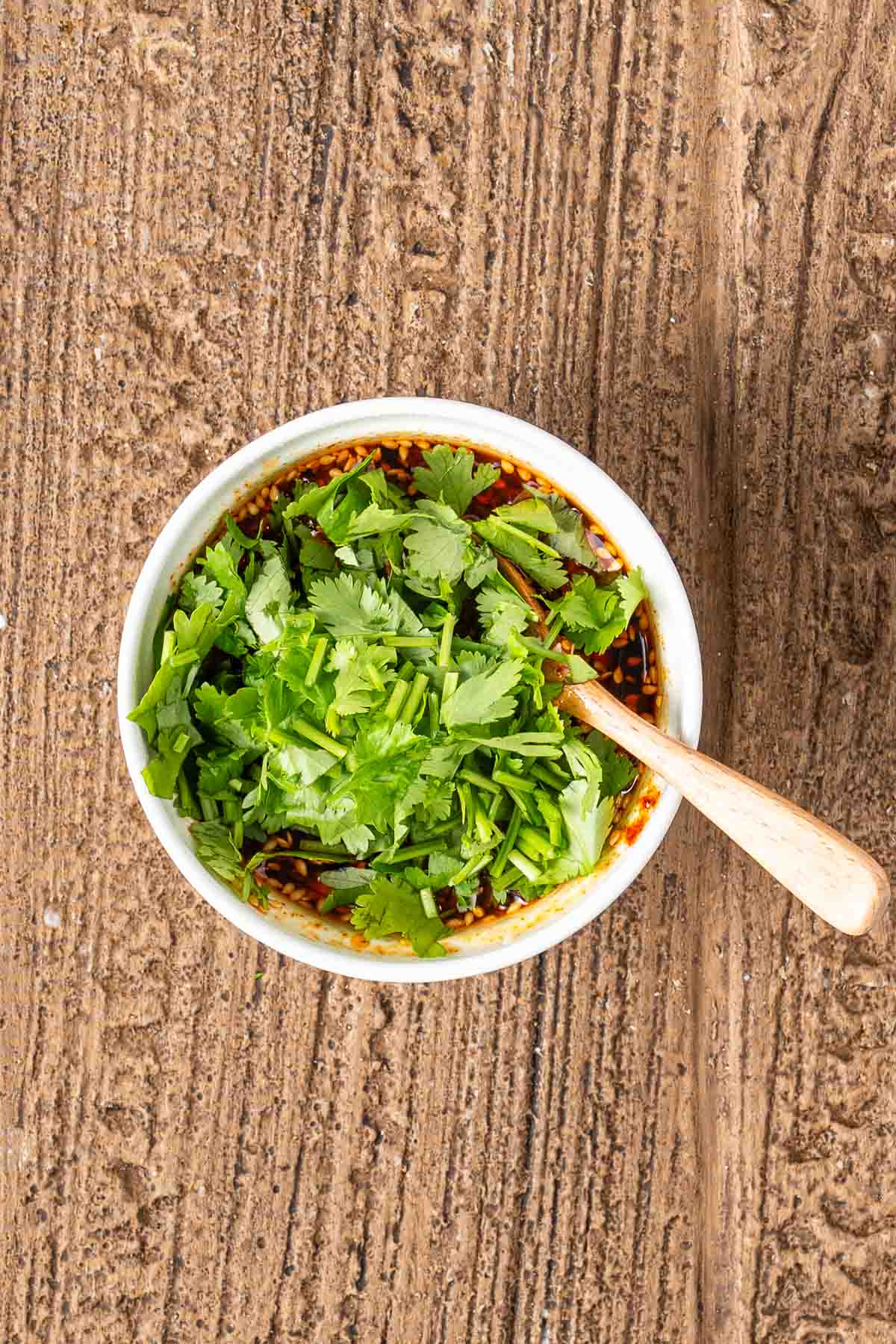 A bowl of soy sauce-based dipping sauce topped with chopped cilantro, with a wooden spoon resting inside, placed on a textured brown surface.