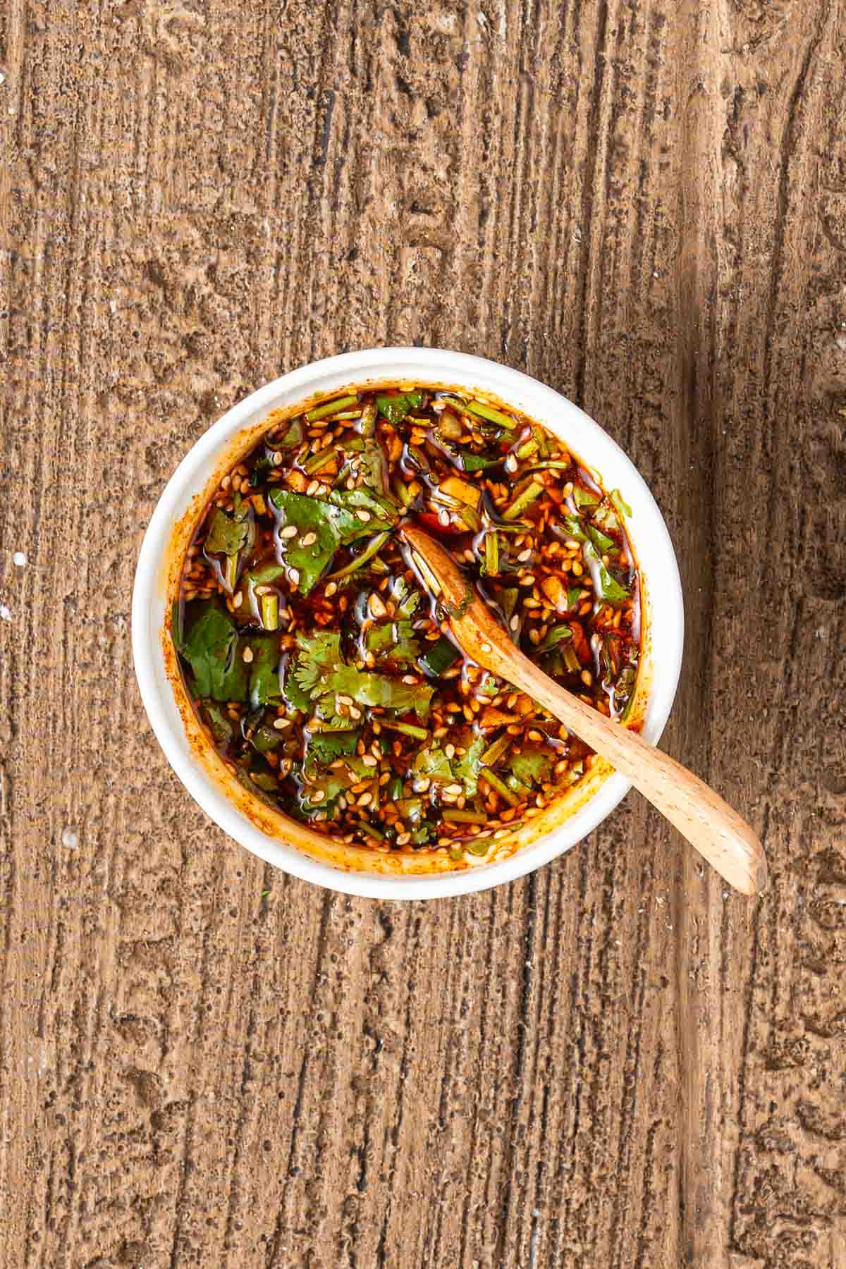 A small white bowl filled with a dark, chunky sauce containing chopped green herbs, sesame seeds, and a wooden spoon, placed on a textured brown surface.