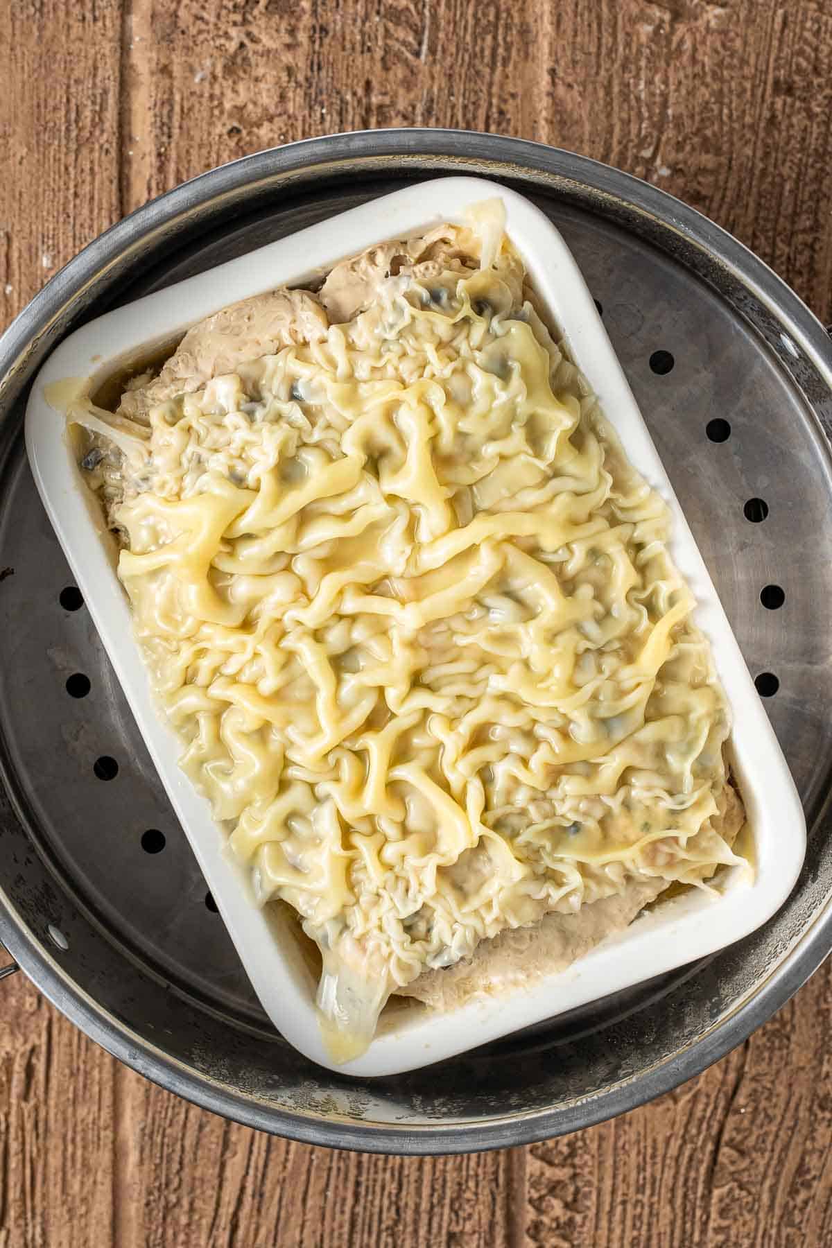 Unbaked lasagna in a white rectangular dish with wavy pasta sheets and cheese on top, placed inside a metal steamer on a wooden surface.