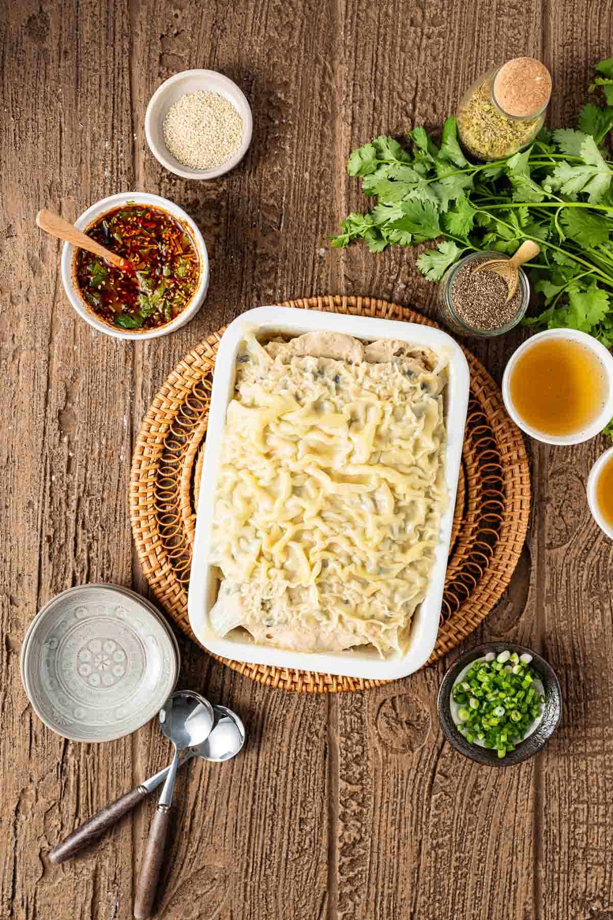 A casserole dish filled with creamy lasagna sits on a woven mat, surrounded by bowls of sauce, herbs, green onions, sesame seeds, and seasonings on a wooden table.