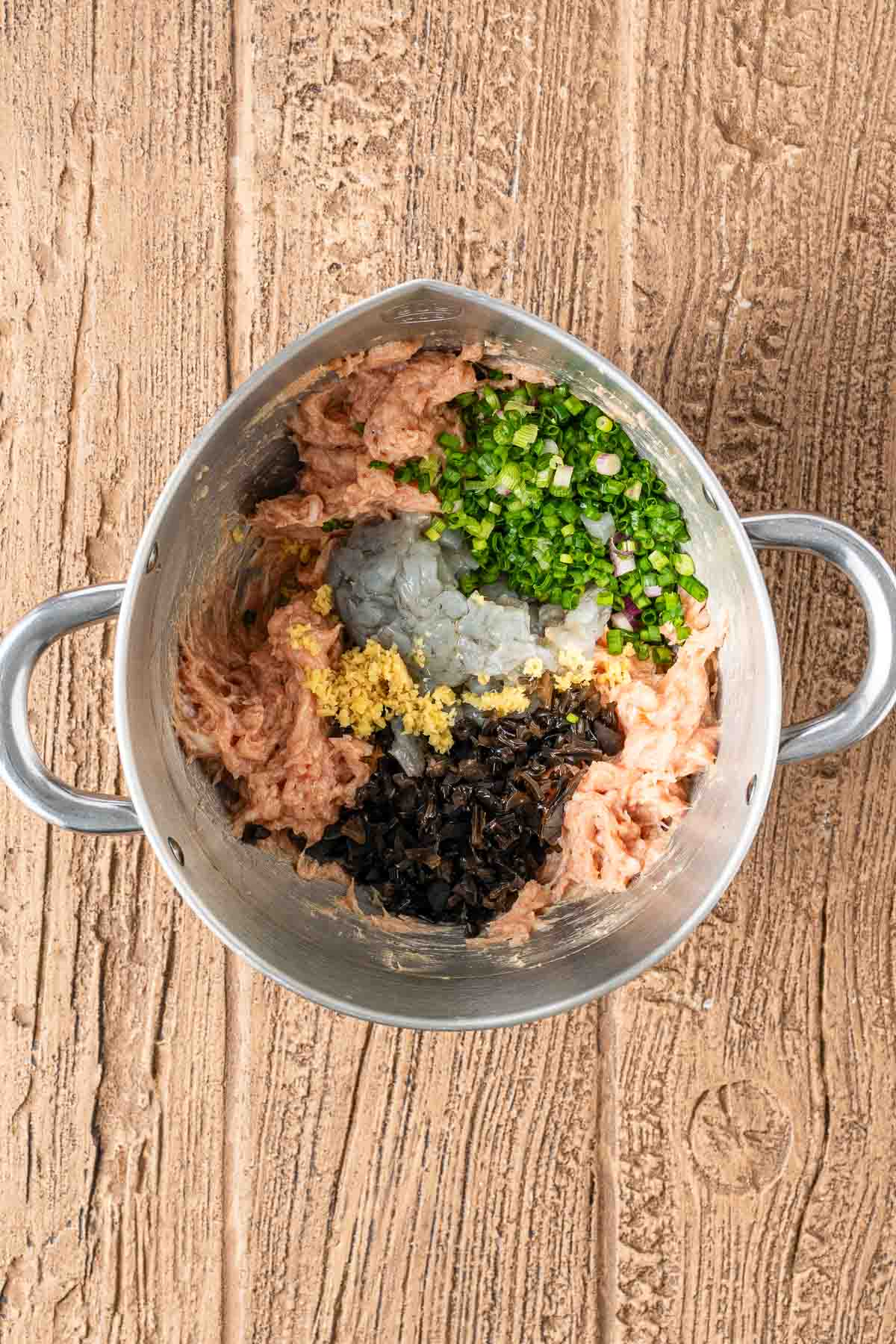 A metal mixing bowl on a wooden surface contains minced meat, chopped green onions, shrimp, ginger, and diced mushrooms, all unmixed.