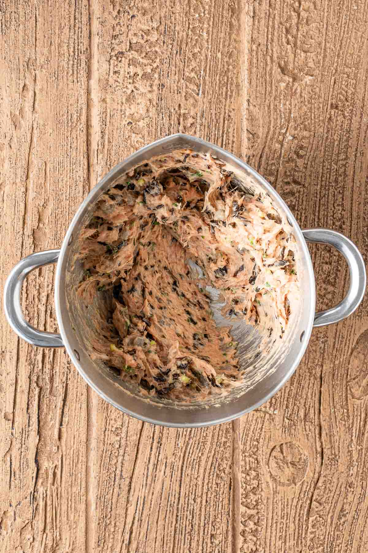 A metal mixing bowl with two handles containing a partially mixed blend of butter and herbs, placed on a wooden surface.