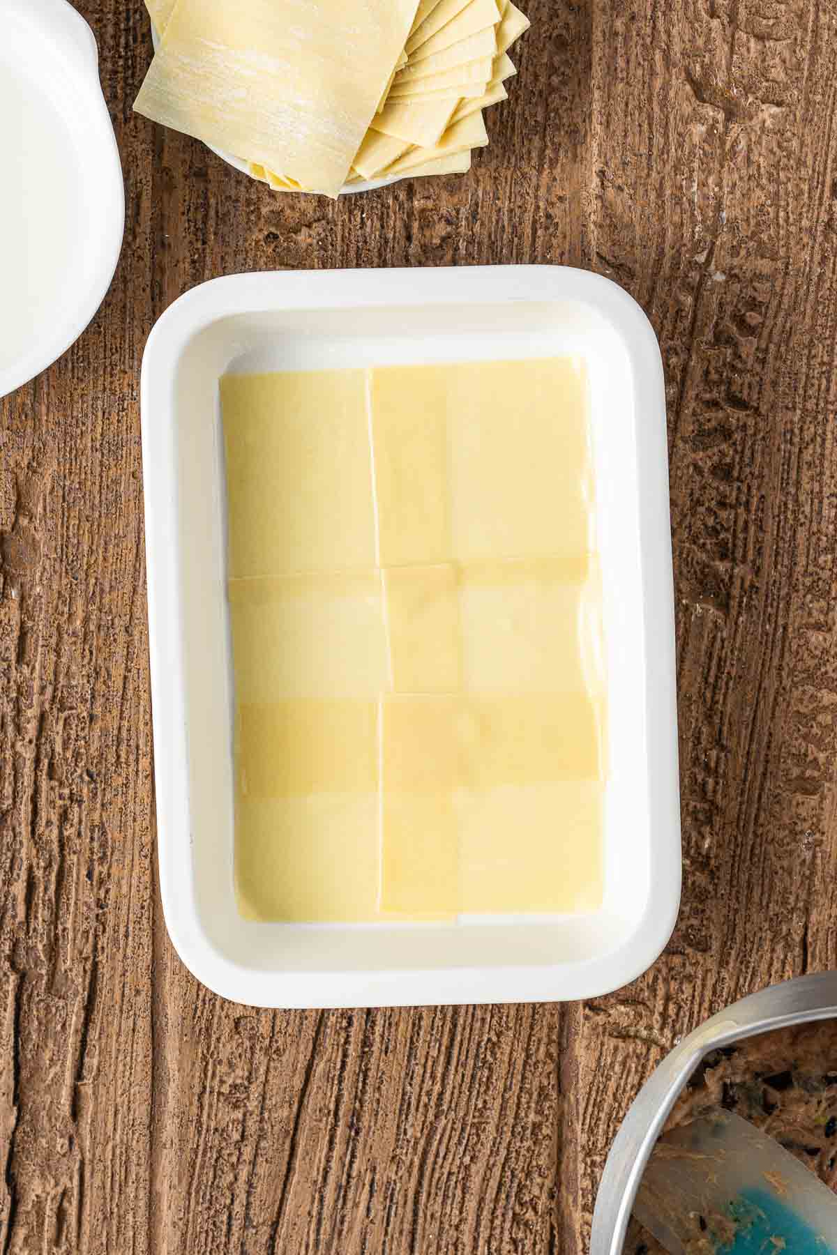 A white rectangular dish with uncooked lasagna noodles arranged in a single layer, on a wooden surface.