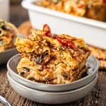 A close-up of a slice of vegetable lasagna topped with herbs and sauce, served on a small plate with a baking dish of lasagna in the background.