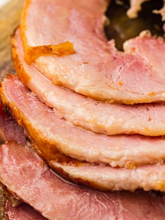 Several slices of cooked ham are arranged in a stack, showing their pink interior and slightly browned edges on a wooden surface.