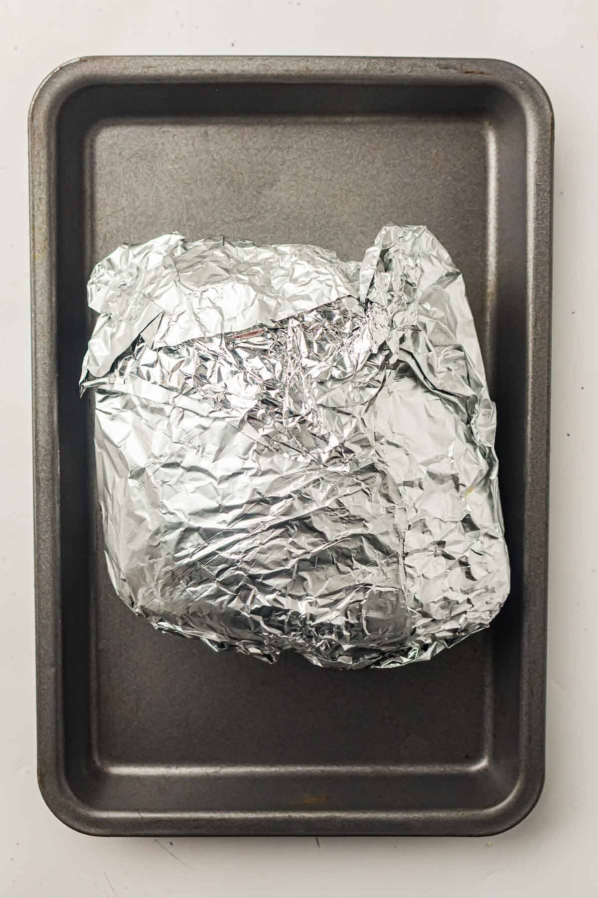 A rectangular baking tray holding a large food item wrapped in crumpled aluminum foil.