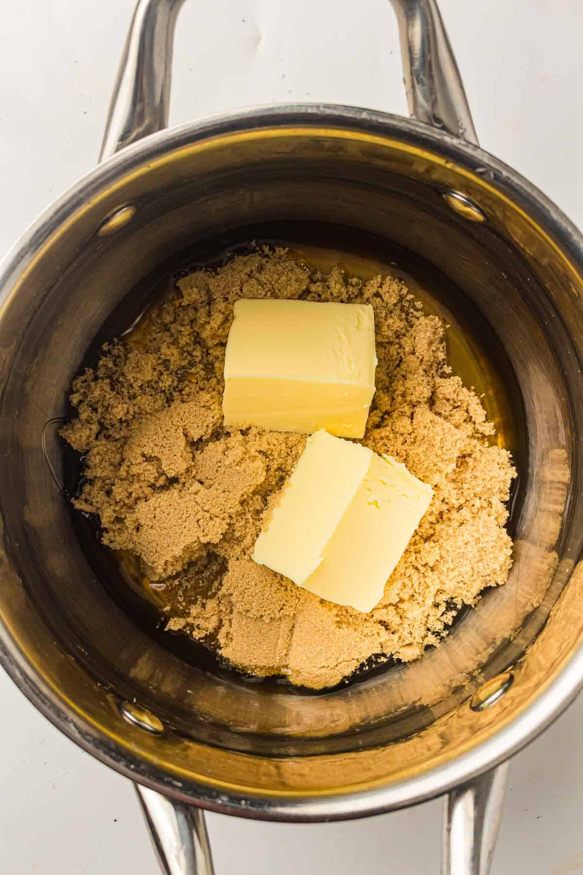 A metal saucepan containing brown sugar and two rectangular pieces of butter, viewed from above.