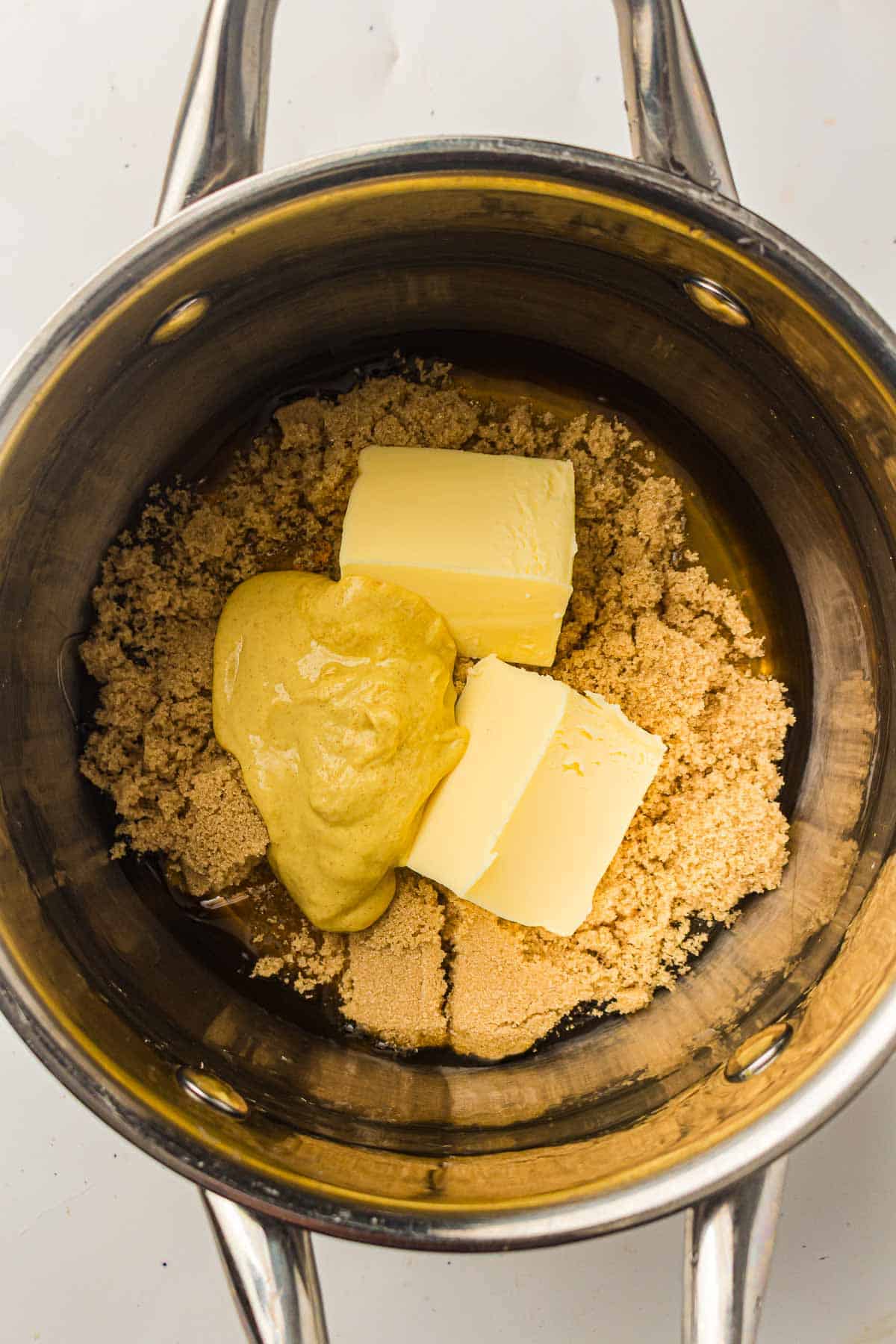 A saucepan containing brown sugar, two slabs of butter, and a dollop of yellow mustard, ready to be mixed.