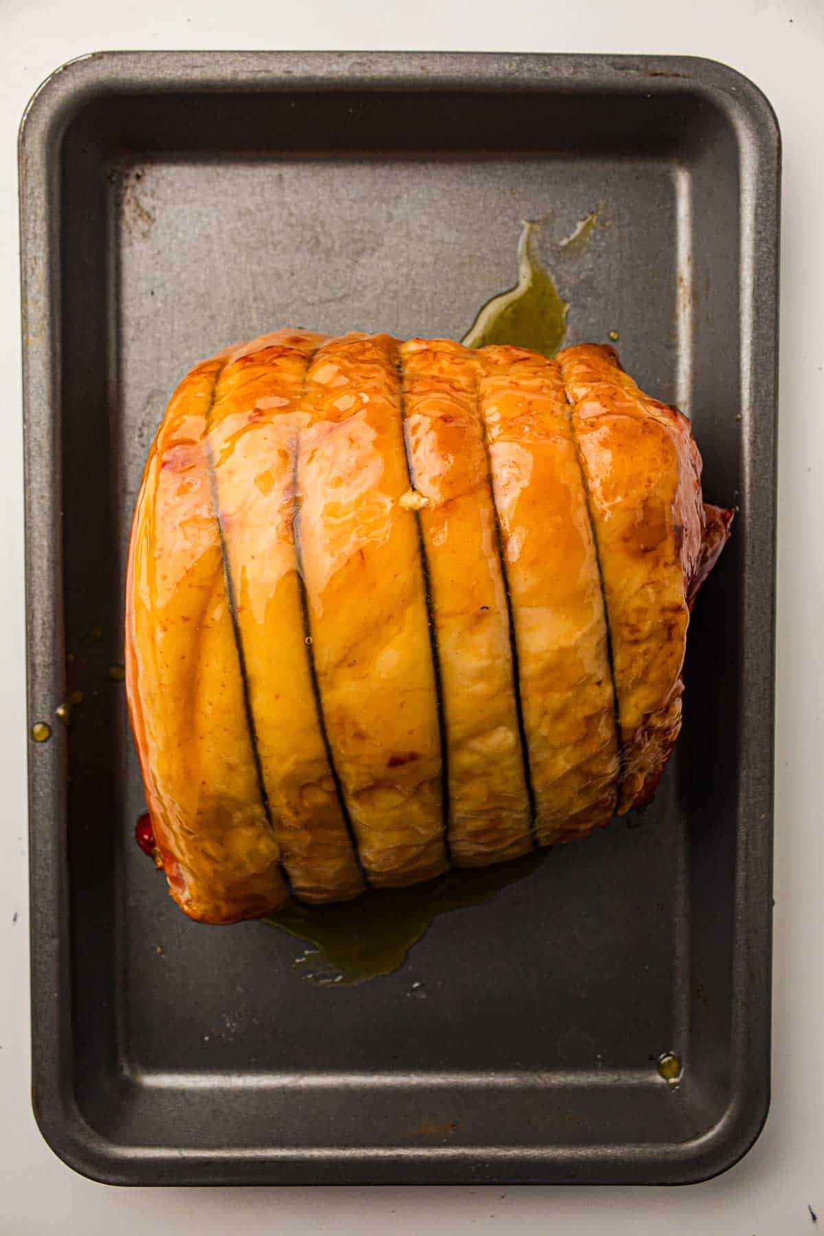 A cooked, glazed ham roast sits in a rectangular metal baking pan, showing scored lines across the top and some juices in the pan.