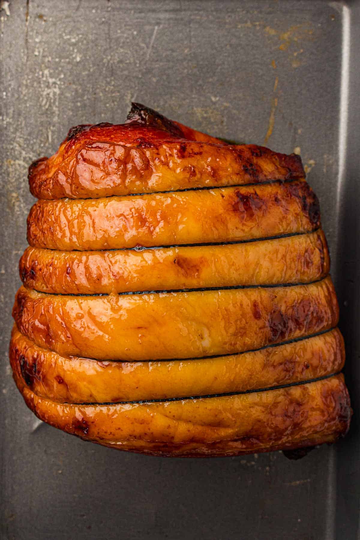 A cooked, sliced, glazed ham sits on a dark metal baking sheet.