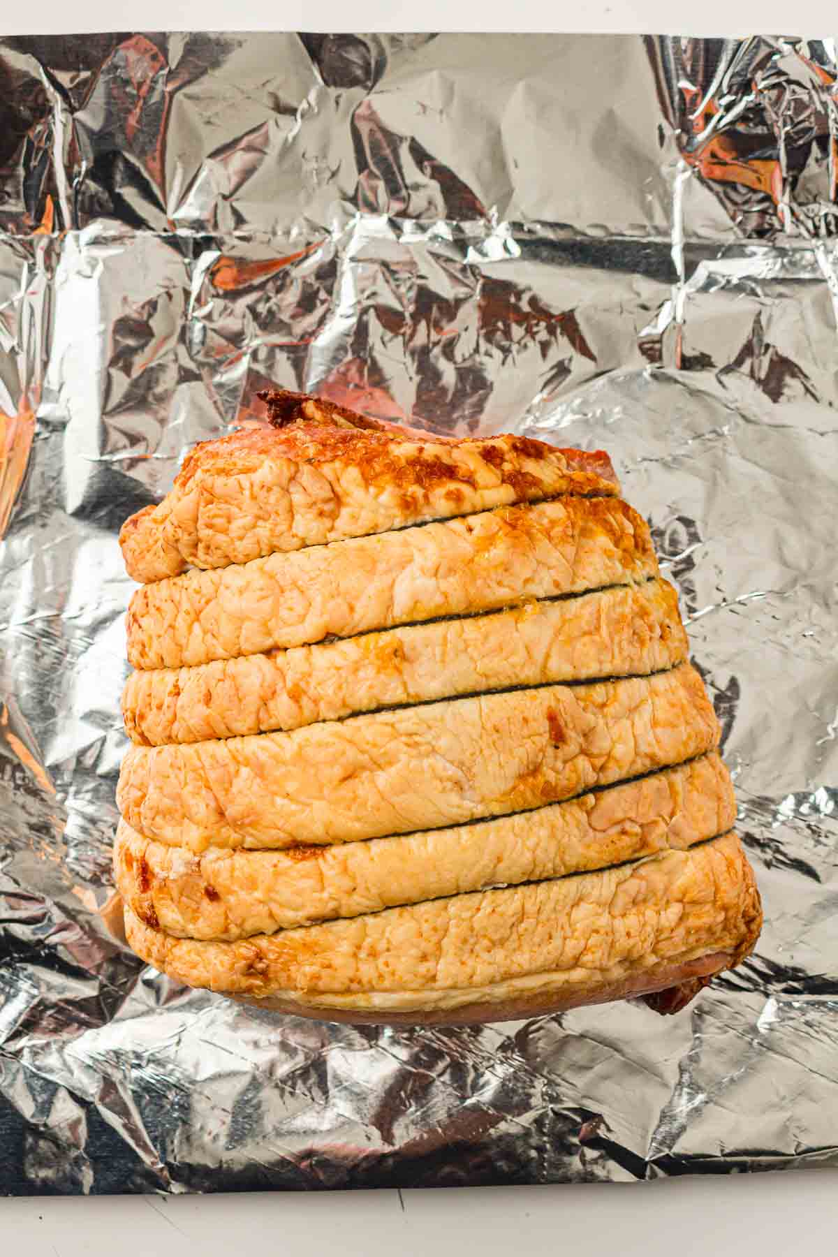 A cooked boneless ham, sliced and tied, sits on a sheet of crumpled aluminum foil.