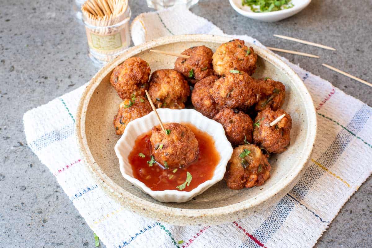 A bowl of fried meatballs with toothpicks, served with a small dish of dipping sauce, placed on a plate atop a white towel on a gray countertop.