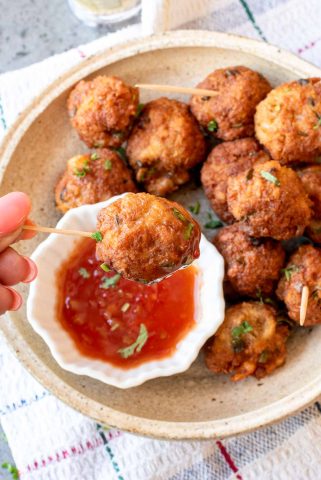 A hand holds a skewer with a fried ball dipped in red sauce; more fried balls are on a beige plate in the background.