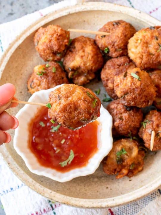 A hand holds a skewer with a fried ball dipped in red sauce; more fried balls are on a beige plate in the background.