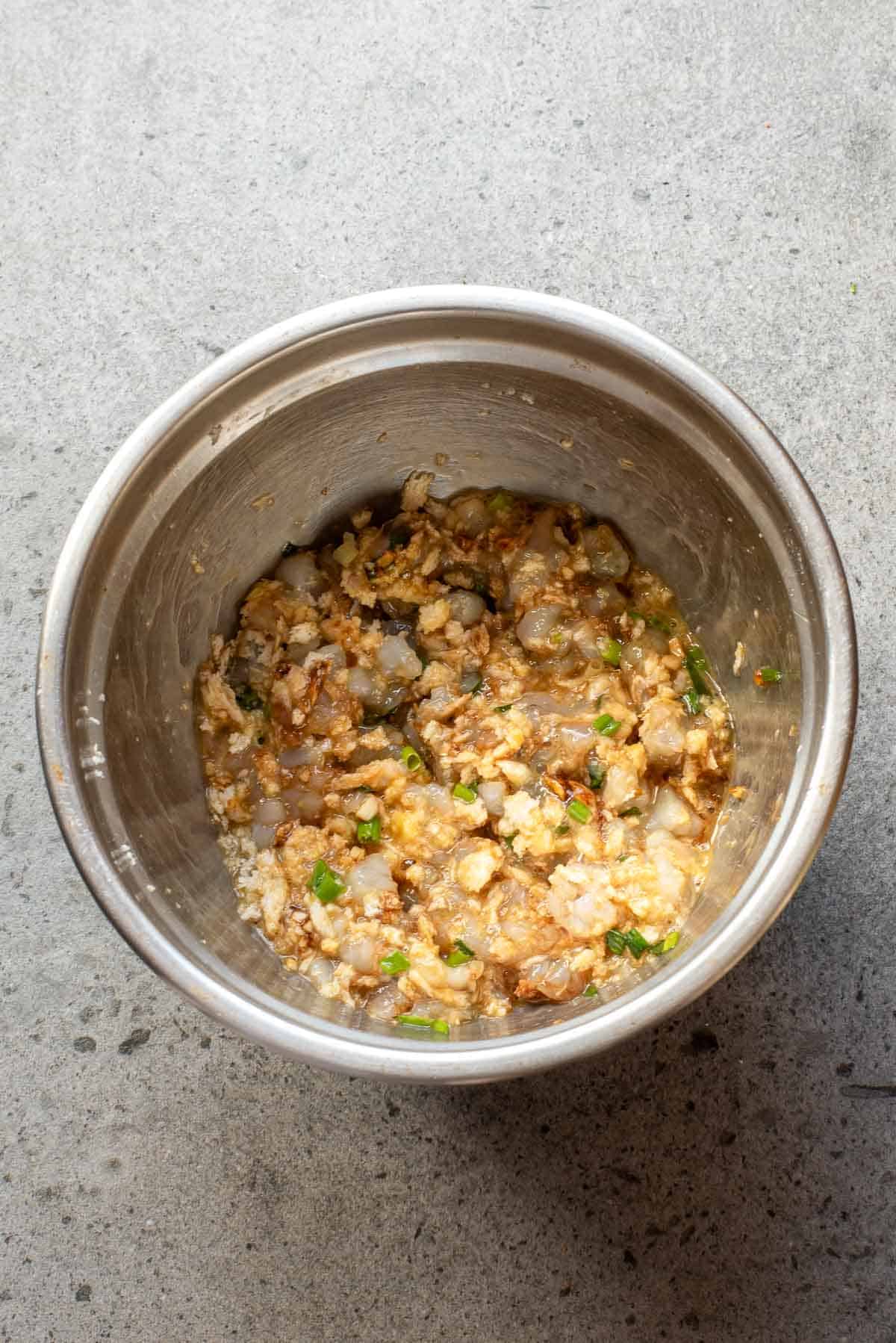 A metal mixing bowl containing a mixture of raw shrimp, ground meat, chopped green onions, and seasonings on a gray surface.