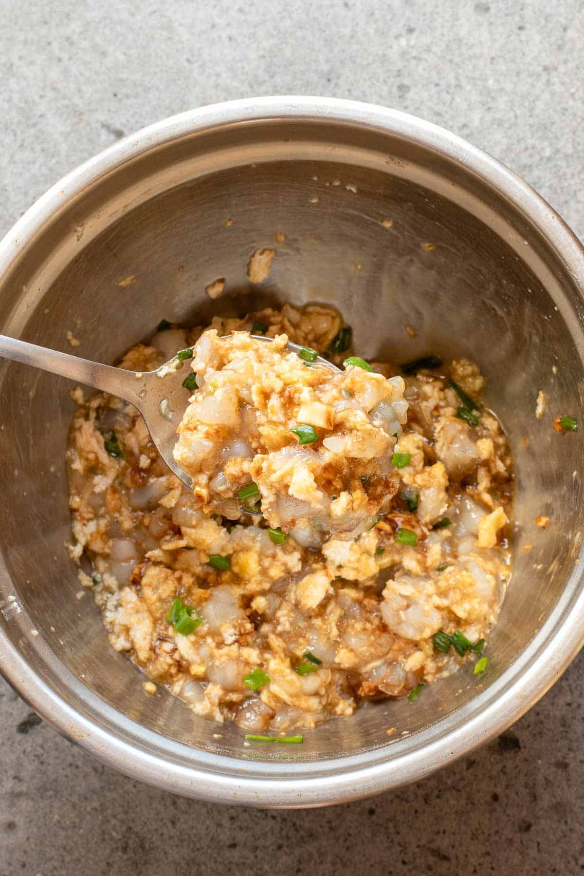 A metal bowl containing a mixture of chopped raw shrimp, egg, and chopped green onions, with a spoon scooping some of the mixture.