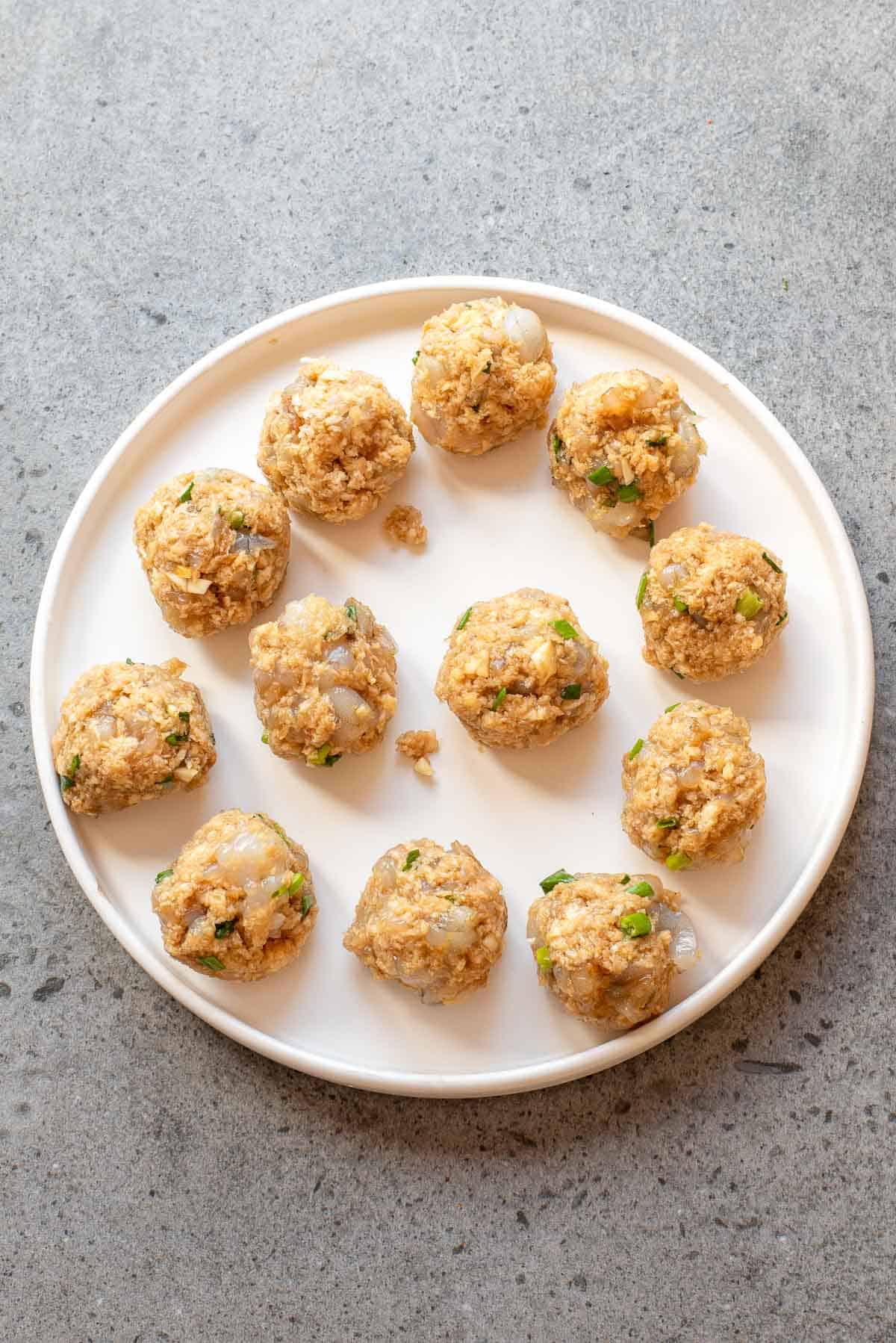 A round white plate holds twelve uncooked shrimp meatballs mixed with herbs on a gray, textured surface.